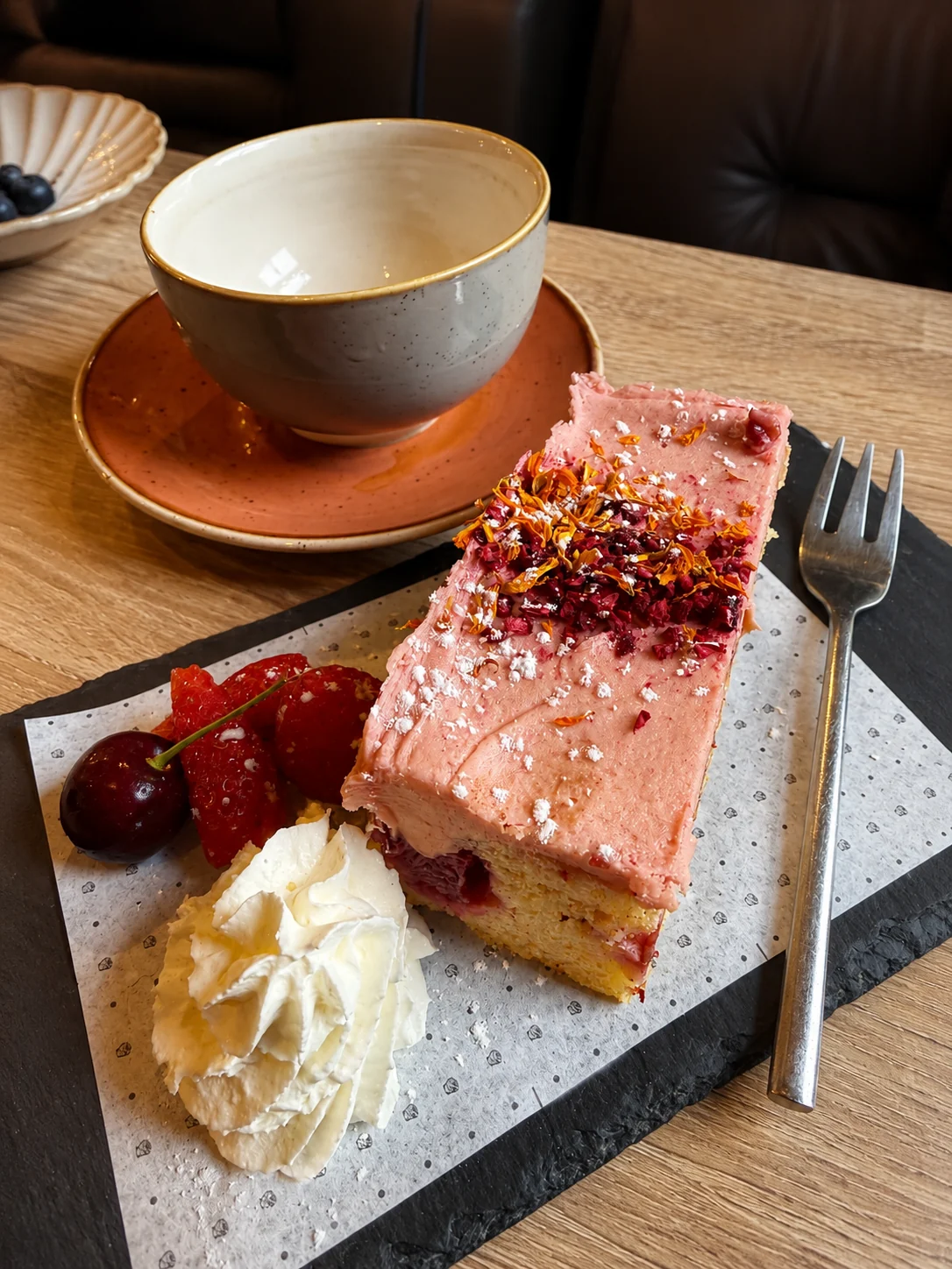 Pink iced cake served with fruit beside a cup